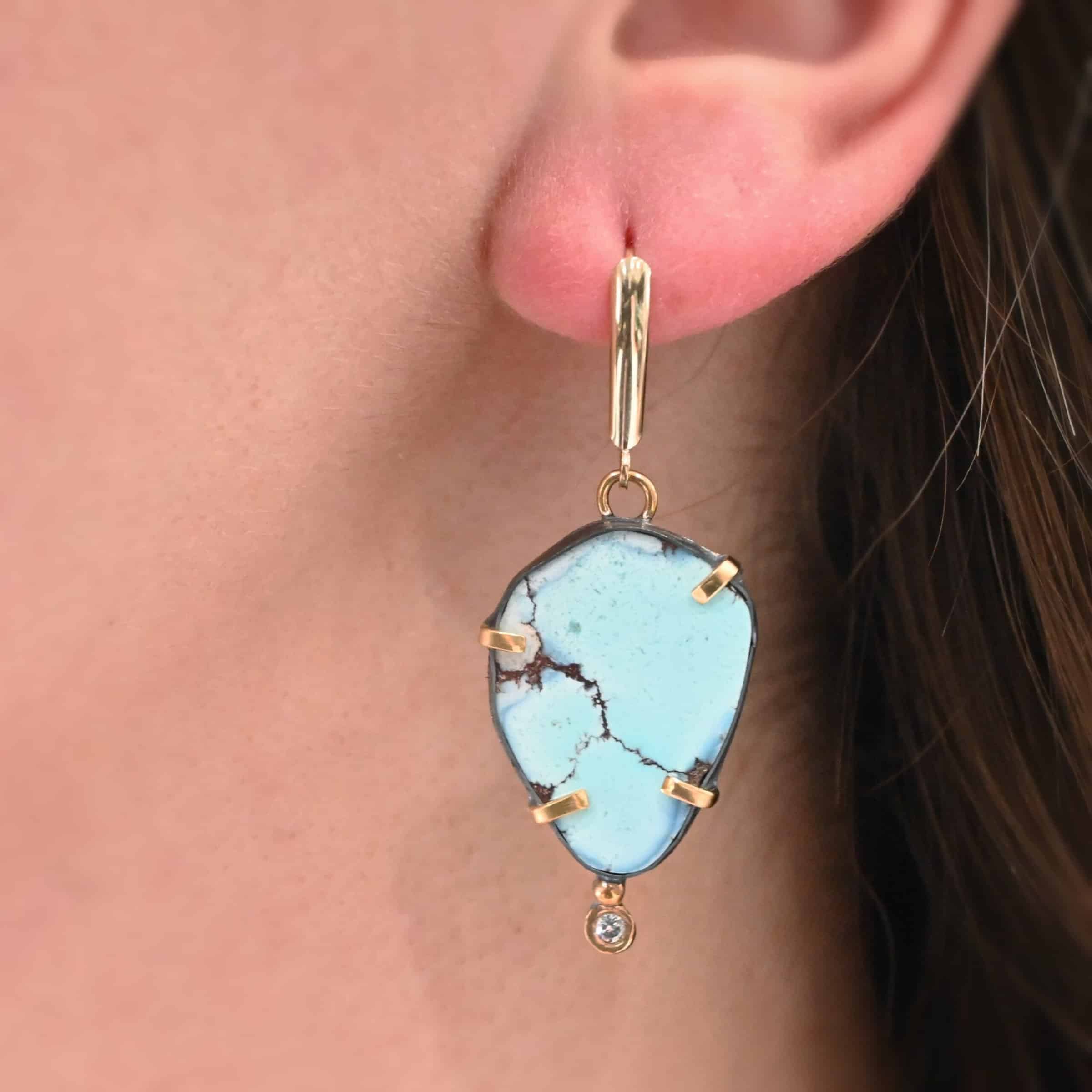 Path Of A Glacier Earrings Photo of one of the "Path Of A Glacier" Earrings. Made out of sterling silver, gold, turquoise, and lab created diamonds. Earrings has a large oval shaped turquoise in the center with four gold prongs, two per side, holding the stones in. There is a gold ball on the bottom turquoise of the right earring and a diamond on the bottom of the gold ball. At the top of the earring, on the back of it is a gold loop. Attached to the loops is a gold leverback ear wire. Earring is worn up close by a model to demonstrate size.