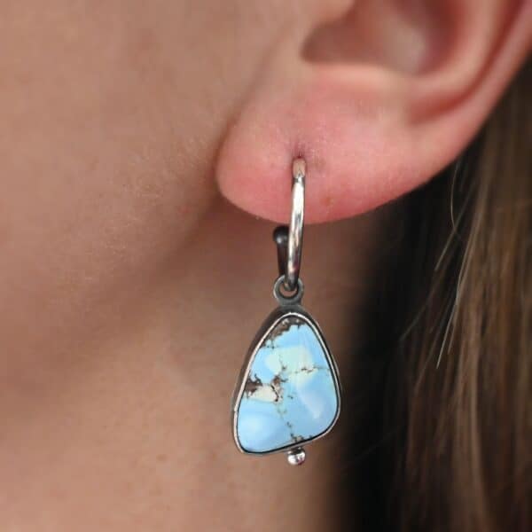 Photo of the "Frozen Land" Earring up close. Made out of sterling silver and turquoise. The turquoise is a triangle shape and there is a silver ball on the bottom of the turquoise stone setting. The turquoise is a mix of light and dark blue, with dark brown lines going through the entire stone. There is a silver loop soldered to the top of the setting. Going through this loop is the half circle post earring. Earring is worn by a model to demonstrate size.