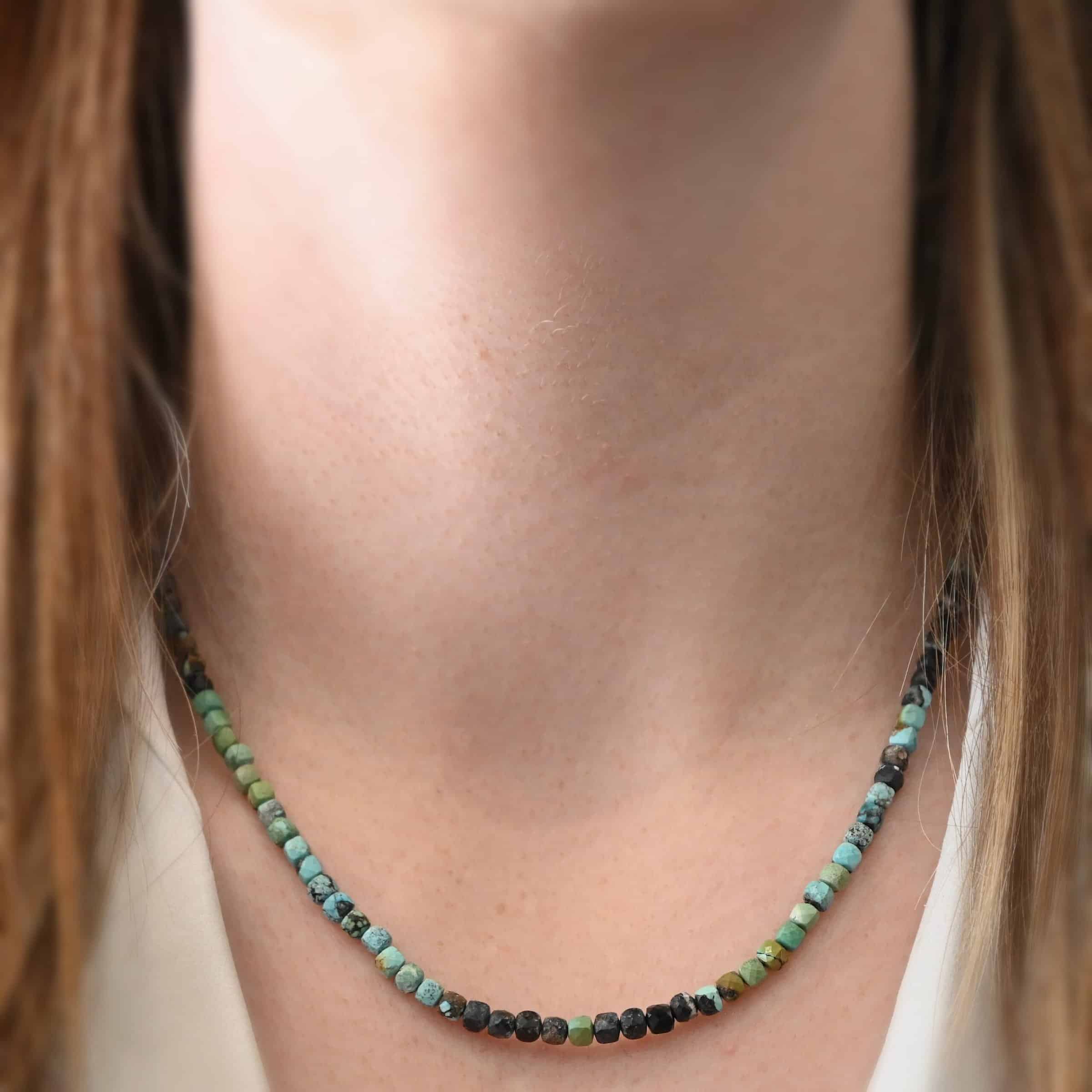 Ada Necklace Close up of the "Ada" Necklace as worn by a model. Necklace is made out of larger sized, square, turquoise beads. The beads are strung on heavy wire and are right next to each other tightly. Beads are a mix of blue, green, black, and brown colors. Necklace is worn at 17" long to demonstrate size.