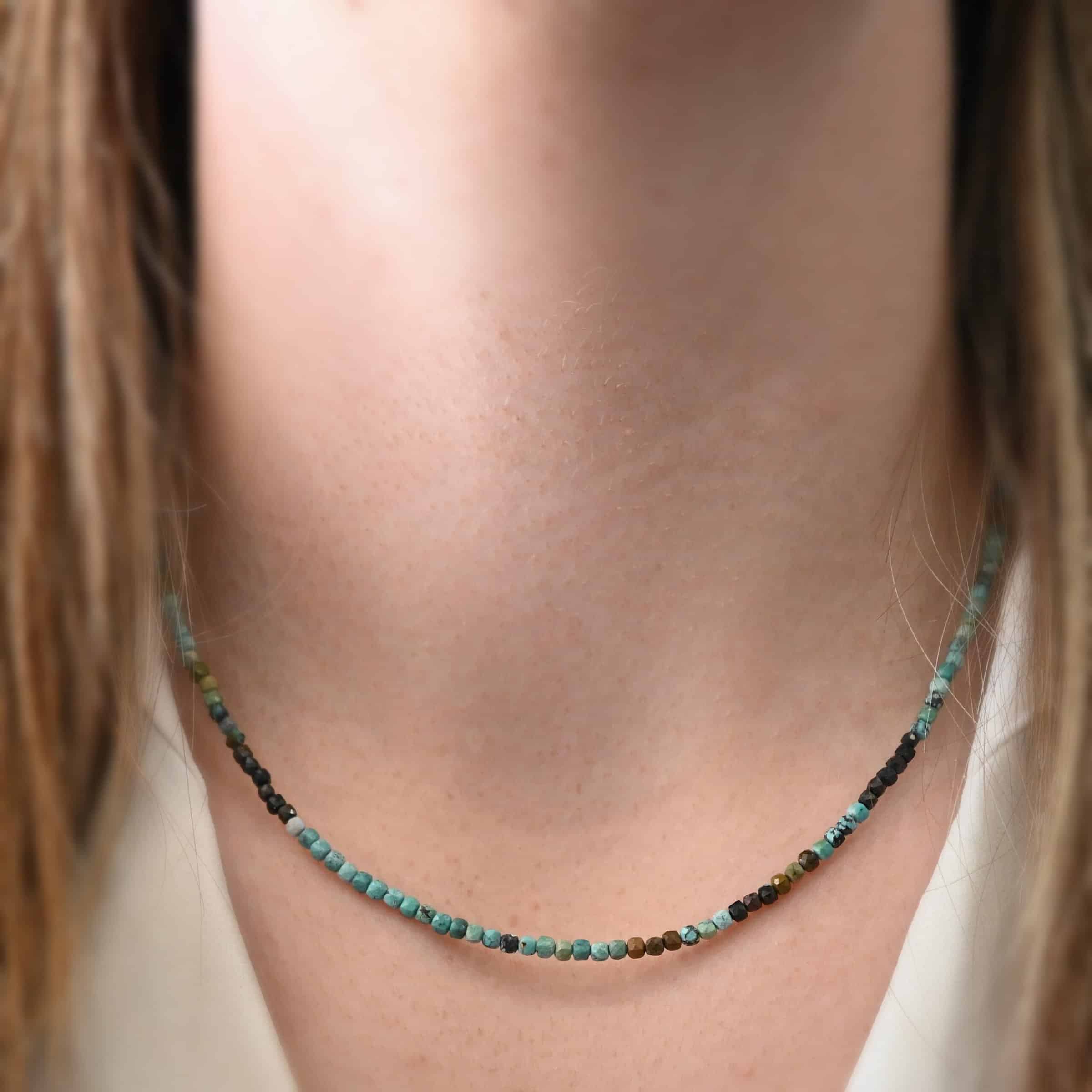 Lenora Necklace Close up of the "Lenora" Necklace as worn by a model. Necklace is made out of smaller sized, square, turquoise beads. The beads are strung on heavy wire and are right next to each other tightly. Beads are a mix of blue, green, black, and brown colors. Necklace is worn at 17" long to demonstrate size.