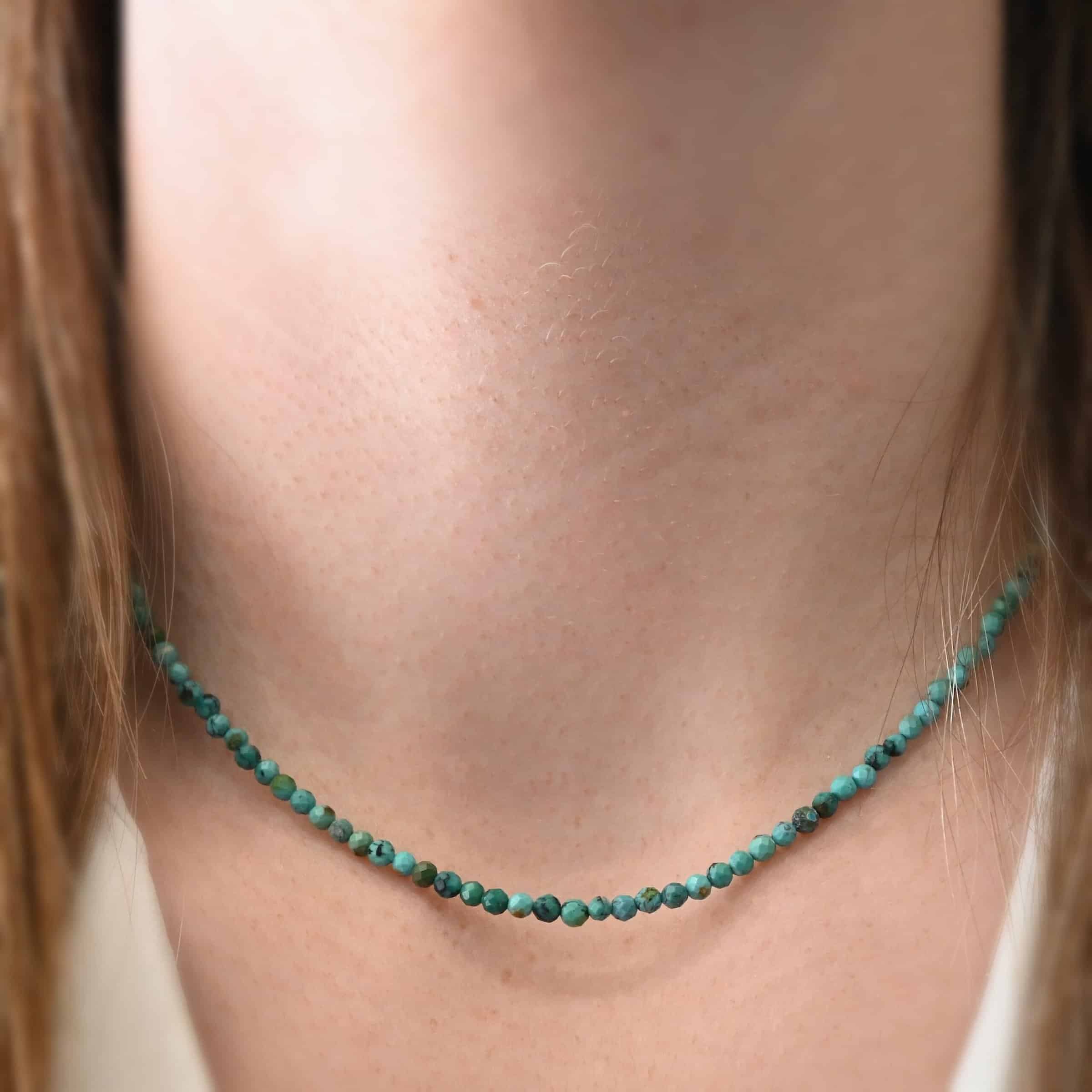 Close up of the "Clara" Necklace as worn by a model. Necklace is made out of smaller sized, spherical, turquoise beads. The beads are strung on heavy wire and are right next to each other tightly. Beads are a mix of blue and green colors. Necklace is worn at 17" long to demonstrate size.