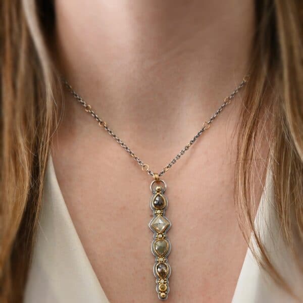 Photo of the "Daybreak" Necklace. Made out of sterling silver, gold, and diamond. There are nine diamonds, all in a straight line, that create the pendant of the necklace. Five of the diamonds are different shapes, sizes, and colors. Each of these diamonds are separated by small, circular diamonds, which there are four of. The top of the pendant starts with a large diamond, then a small one, and keeps alternating until the final diamond is a large one. There is a large loop soldered to the back of the top of the pendant with four gold jump rings attached to it. Going through the jump rings is the silver chain of the necklace. The chain of the necklace has gold loops scattered around it. Necklace is worn by a model at 16" long to demonstrate size of the pendant.