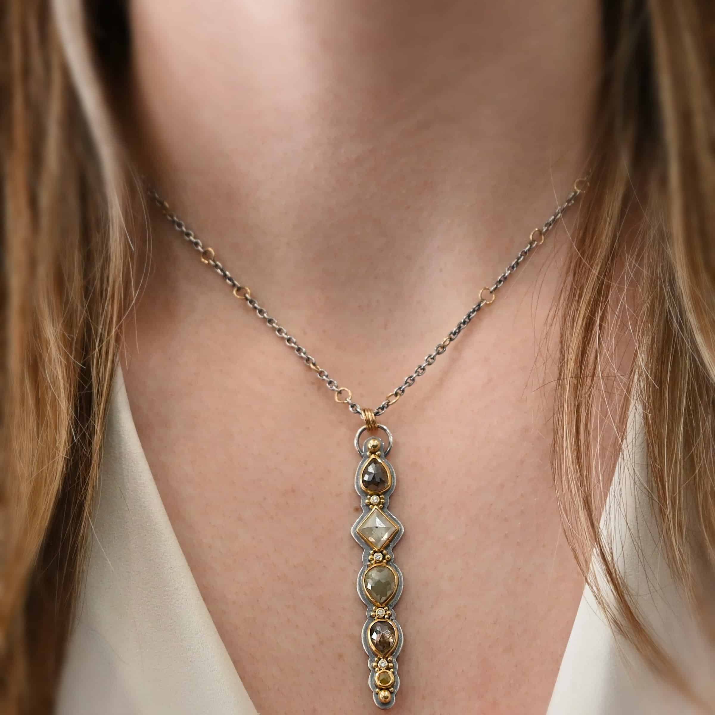 Daybreak Necklace Photo of the "Daybreak" Necklace. Made out of sterling silver, gold, and diamond. There are nine diamonds, all in a straight line, that create the pendant of the necklace. Five of the diamonds are different shapes, sizes, and colors. Each of these diamonds are separated by small, circular diamonds, which there are four of. The top of the pendant starts with a large diamond, then a small one, and keeps alternating until the final diamond is a large one. There is a large loop soldered to the back of the top of the pendant with four gold jump rings attached to it. Going through the jump rings is the silver chain of the necklace. The chain of the necklace has gold loops scattered around it. Necklace is worn by a model at 16" long to demonstrate size of the pendant.