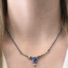 Photo of the "All In Harmony" Necklace. Made out of Sterling Silver, Gold, Tanzanite, Sapphires, Tourmaline, Amethyst, & Diamond. The center of the pendant is a pear shaped tanzanite, with the point facing down. There is a small diamond beneath the tanzanite, on the tip of the stone. On each side of the tanzanite, on the bar of the necklace that the top of the tanzanite is soldered to, are four stones, two per side. The left side on the bar has a green tourmaline and a yellow sapphire. The right side has a pink sapphire and an amethyst. There are multiple gold balls scattered around the stones on the top. There is also a thin line of gold that goes across the curved bar. Necklace is worn by a model at 17" long to demonstrate size of the necklace.