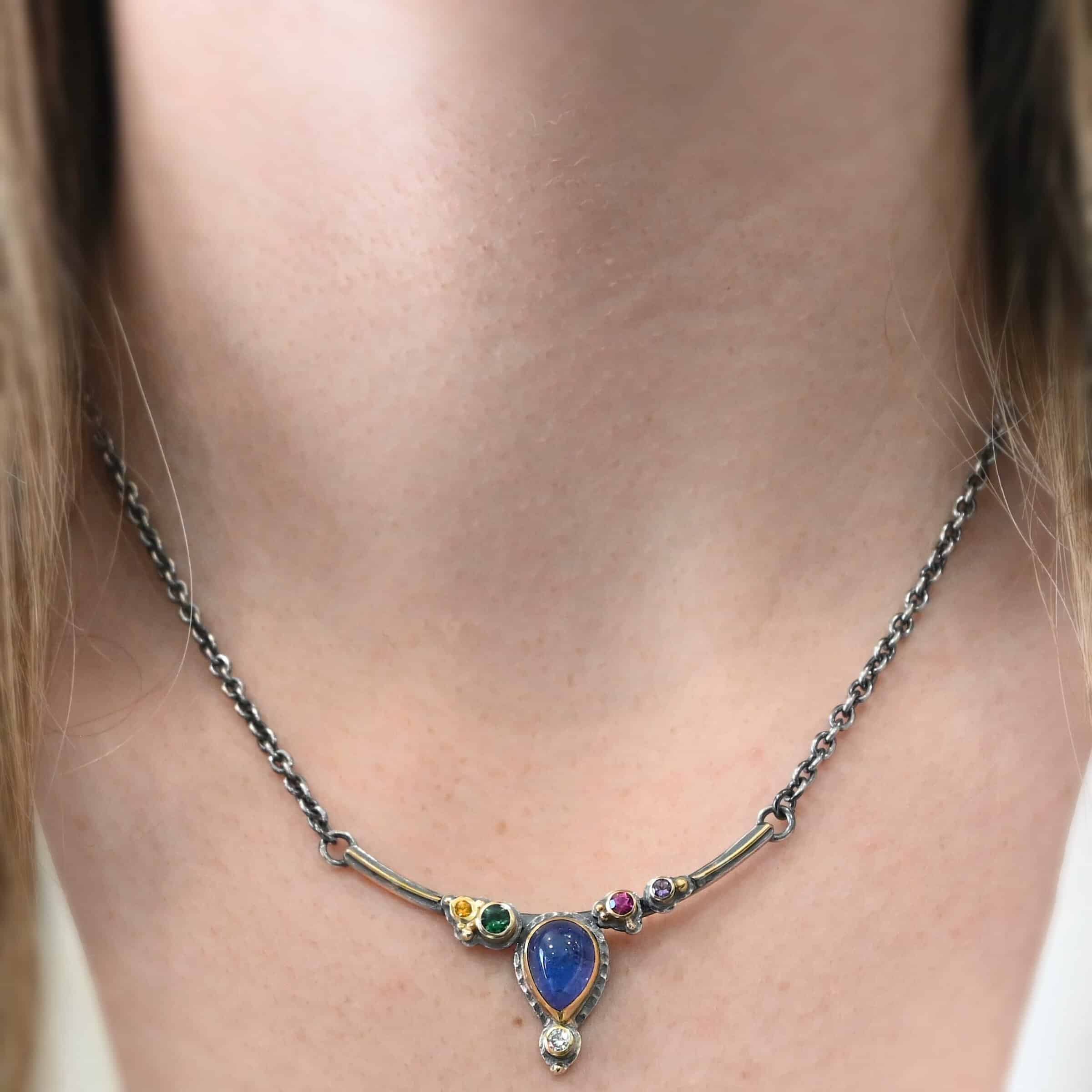 Photo of the "All In Harmony" Necklace. Made out of Sterling Silver, Gold, Tanzanite, Sapphires, Tourmaline, Amethyst, & Diamond. The center of the pendant is a pear shaped tanzanite, with the point facing down. There is a small diamond beneath the tanzanite, on the tip of the stone. On each side of the tanzanite, on the bar of the necklace that the top of the tanzanite is soldered to, are four stones, two per side. The left side on the bar has a green tourmaline and a yellow sapphire. The right side has a pink sapphire and an amethyst. There are multiple gold balls scattered around the stones on the top. There is also a thin line of gold that goes across the curved bar. Necklace is worn by a model at 17" long to demonstrate size of the necklace.