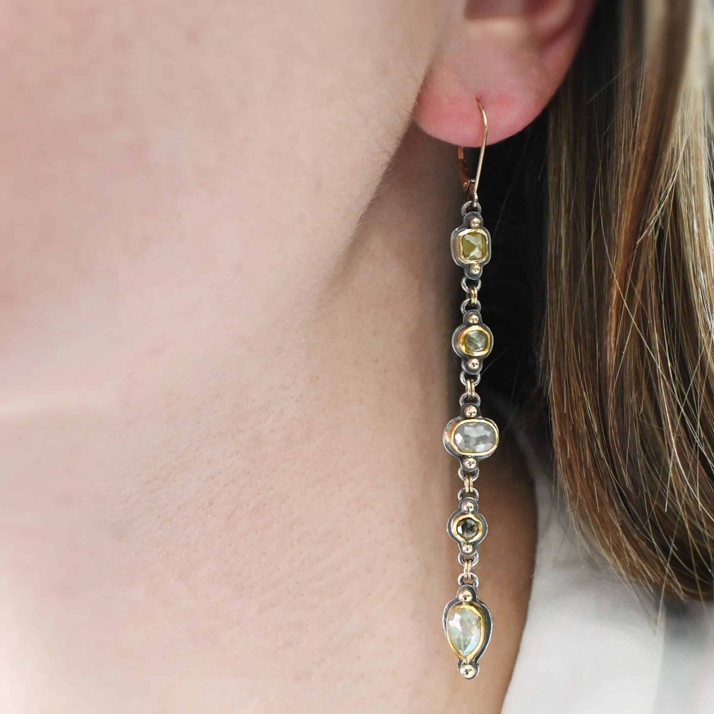 Photo of the "First Light" Earrings. Made out of sterling silver, gold, and diamonds. The top of the earrings are gold leverback ear wires that are connected to small silver loops on the back of the top of the earrings. Each earring has five diamonds that all hang down. Each diamond is set in gold, and the back of each diamond has a loop on the top and bottom to connect it to the previous diamond and the next diamond. Each diamond has two gold balls on the front, one on the top and one on the bottom. Each diamond is connected by two gold jump rings that go through the loops on the back. Earrings are worn up close by a model.