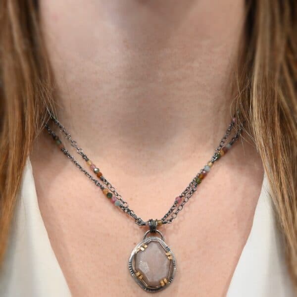 Photo of the "Peach Tree" Necklace. Made out of sterling silver, gold, peach moonstone, and tourmaline. There is a rose cut, octagon shaped peach moonstone in the center with eight gold prongs holding the stone in. The prongs are four groups of two in each corner of the ring. The silver around the stone has hammered marks in groups scattered around it as well. There is a loop on the back of the pendant with three silver jump rings going through it. Going through the jump rings is the chain of the necklace. The chain of the necklace is two separate chains that meet at the back to make one chain for the necklace. Each chain has multiple tourmaline beads on it. Necklace is worn by a model at 17" long to demonstrate size and how the necklace lies down when it is worn.
