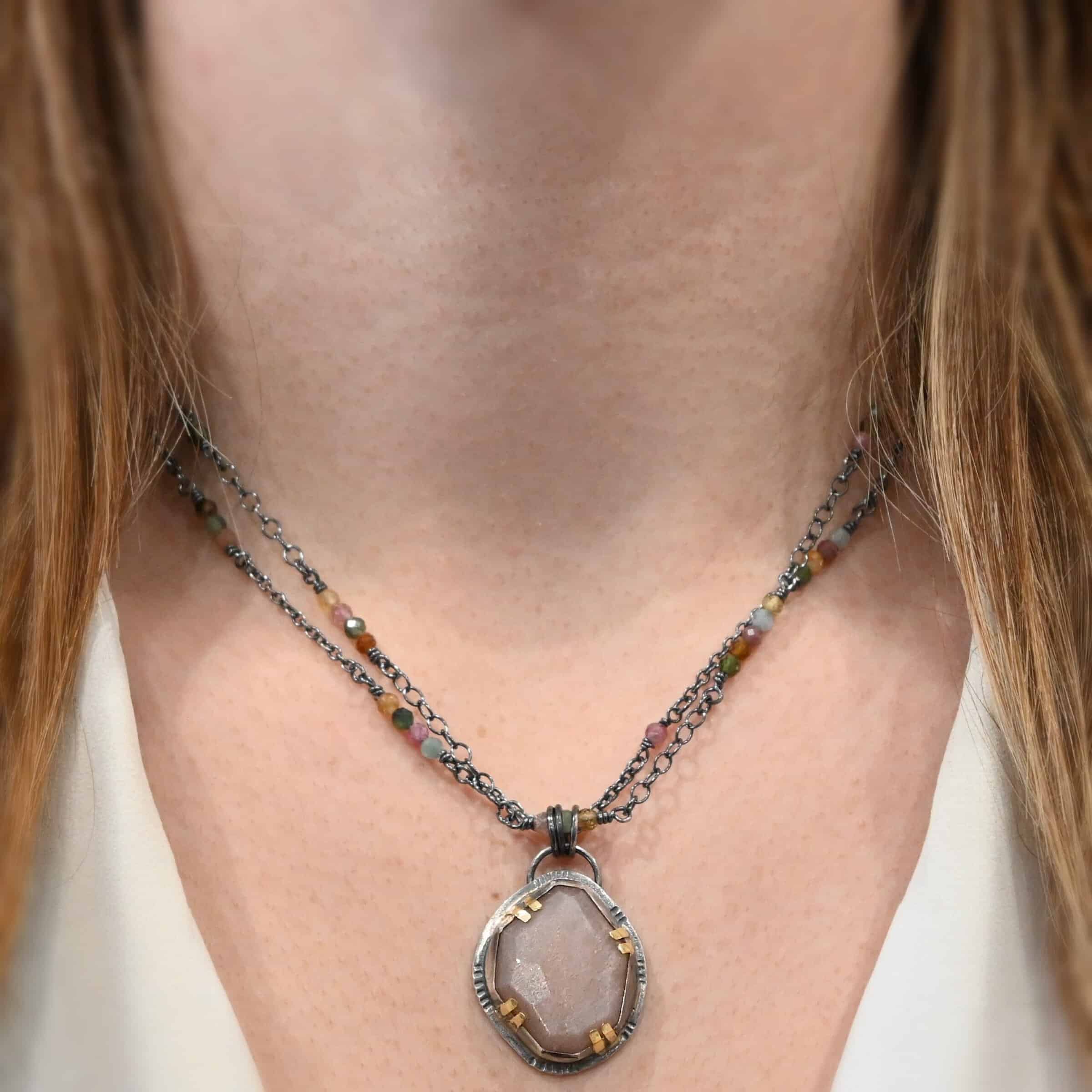 Peach Tree Necklace Photo of the "Peach Tree" Necklace. Made out of sterling silver, gold, peach moonstone, and tourmaline. There is a rose cut, octagon shaped peach moonstone in the center with eight gold prongs holding the stone in. The prongs are four groups of two in each corner of the ring. The silver around the stone has hammered marks in groups scattered around it as well. There is a loop on the back of the pendant with three silver jump rings going through it. Going through the jump rings is the chain of the necklace. The chain of the necklace is two separate chains that meet at the back to make one chain for the necklace. Each chain has multiple tourmaline beads on it. Necklace is worn by a model at 17" long to demonstrate size and how the necklace lies down when it is worn.