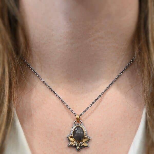 Photo of the "Golden Fields" Necklace. Made out of sterling silver, gold, sapphire, and diamonds. The center stone is an oval shaped, deep brown sapphire that is set in gold. There is a gold ball on the left and right side of this sapphire. There are also four gold leaves, two on each side, beneath the gold balls. The ends of each leaf meet, and where they meet in the center is a gold ball. There are three diamonds, two small and one big, beneath the sapphire, that the gold leaves surround. The two small diamonds are on the side with the big one in the middle. There are three more gold balls that surround each side of the big diamond. There is a loop on the back of the necklace, with three gold jump rings that are attached to it. Going through the jump rings is the chain of the necklace. Necklace is worn by a model at 17" long to demonstrate size of the necklace.