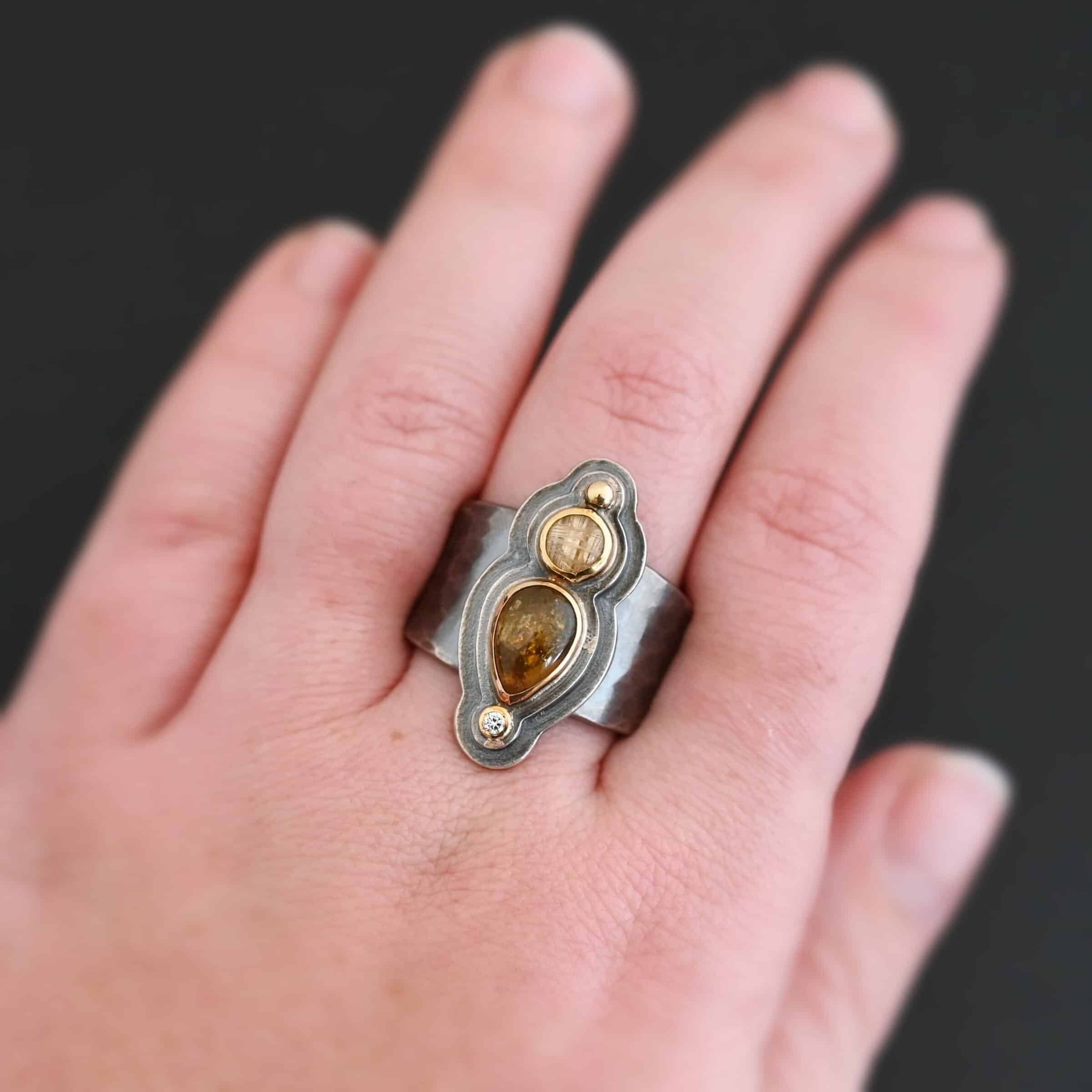 Reach For The Sun Ring Photo of the "Reach For The Sun" Ring. Made out of sterling silver, gold, tourmaline, rutilated quartz, and lab created diamond. The lab created diamond is at the top, on the tip of the pear shaped tourmaline. The tourmaline is a dark brown color with golden flecks. There is a small circular rutilated quartz beneath the tourmaline. On the bottom of the rutilated quartz is a gold ball. Each stone is set in gold, and there are two layers of sterling silver beneath the stones. The top layer is smaller than the bottom layer, so that the bottom layer sticks out. The band of the ring is thick and hammered and oxidized. Ring is shown on a models middle finger to demonstrate size.