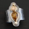 Photo of the "Reach For The Sun" Ring. Made out of sterling silver, gold, tourmaline, rutilated quartz, and lab created diamond. The lab created diamond is at the top, on the tip of the pear shaped tourmaline. The tourmaline is a dark brown color with golden flecks. There is a small circular rutilated quartz beneath the tourmaline. On the bottom of the rutilated quartz is a gold ball. Each stone is set in gold, and there are two layers of sterling silver beneath the stones. The top layer is smaller than the bottom layer, so that the bottom layer sticks out. The band of the ring is thick and hammered and oxidized. Ring is sitting against a dark background.