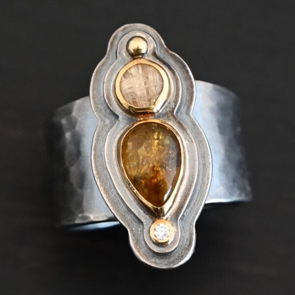 Photo of the "Reach For The Sun" Ring. Made out of sterling silver, gold, tourmaline, rutilated quartz, and lab created diamond. The lab created diamond is at the top, on the tip of the pear shaped tourmaline. The tourmaline is a dark brown color with golden flecks. There is a small circular rutilated quartz beneath the tourmaline. On the bottom of the rutilated quartz is a gold ball. Each stone is set in gold, and there are two layers of sterling silver beneath the stones. The top layer is smaller than the bottom layer, so that the bottom layer sticks out. The band of the ring is thick and hammered and oxidized. Ring is sitting against a dark background.