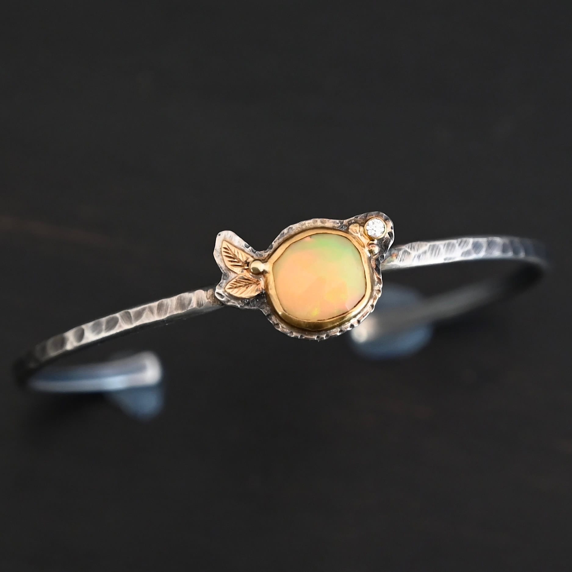 Eternity's Sunrise Cuff Photo of "Eternity's Sunrise" Cuff. Made out of sterling silver, gold, opal, and diamond. There is a circular opal in the center of the cuff. It has vibrant, but translucent red colors shining through it. There is also a light translucent green shining through it. The opal is set in gold. There are two gold leaves on the left side of the cuff, right next to each other. There is a gold dot in the center of the leaves, attached to the gold setting around the opal. There is a diamond set in gold on the right side of the opal. There are two gold dots, one on each side of the diamond. The cuff of the bracelet is made out of sterling silver, and is hammered. Bracelet is sitting against a dark background.