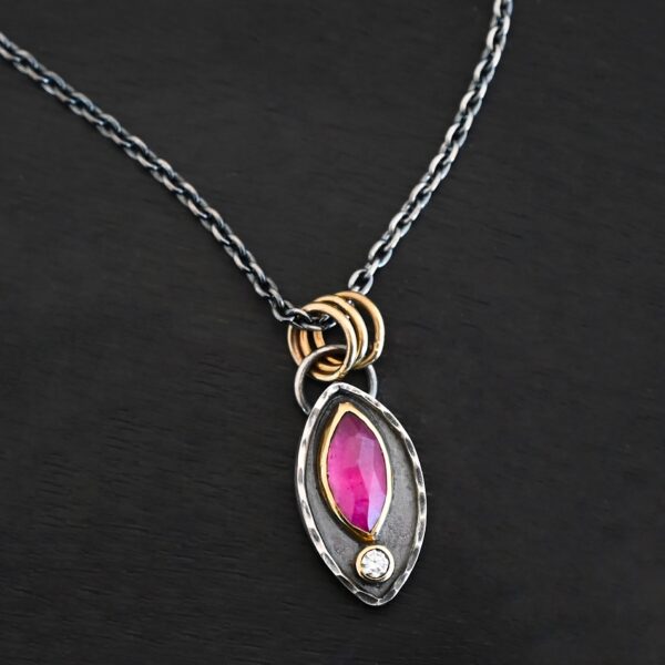 Photo of the "Soft Petals" Necklace. Made out of sterling silver, gold, ruby, and diamond. The bottom of the ring has a small circle diamond. Above this is a pink, marquise shaped ruby placed vertically, so that the points are pointing up and down. The stone is set in gold, and there are two layers of sterling silver beneath it. The top layer of silver goes around the edge of the bottom layer. Both layers are marquise shaped, and the top outside layer is hammered. There is a loop on the back of the necklace in which three gold jump rings go through. Going through the jump rings is the silver chain of the necklace. Necklace is laying against a dark background.