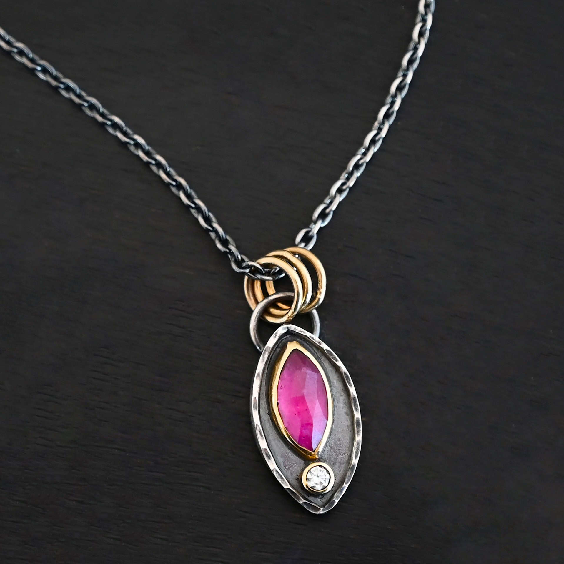 Soft Petals Necklace New Photo of the "Soft Petals" Necklace. Made out of sterling silver, gold, ruby, and diamond. The bottom of the ring has a small circle diamond. Above this is a pink, marquise shaped ruby placed vertically, so that the points are pointing up and down. The stone is set in gold, and there are two layers of sterling silver beneath it. The top layer of silver goes around the edge of the bottom layer. Both layers are marquise shaped, and the top outside layer is hammered. There is a loop on the back of the necklace in which three gold jump rings go through. Going through the jump rings is the silver chain of the necklace. Necklace is laying against a dark background.