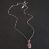 Photo of the "Soft Petals" Necklace. Made out of sterling silver, gold, ruby, and diamond. The bottom of the ring has a small circle diamond. Above this is a pink, marquise shaped ruby placed vertically, so that the points are pointing up and down. The stone is set in gold, and there are two layers of sterling silver beneath it. The top layer of silver goes around the edge of the bottom layer. Both layers are marquise shaped, and the top outside layer is hammered. There is a loop on the back of the necklace in which three gold jump rings go through. Going through the jump rings is the silver chain of the necklace. Necklace is laying against a dark background.