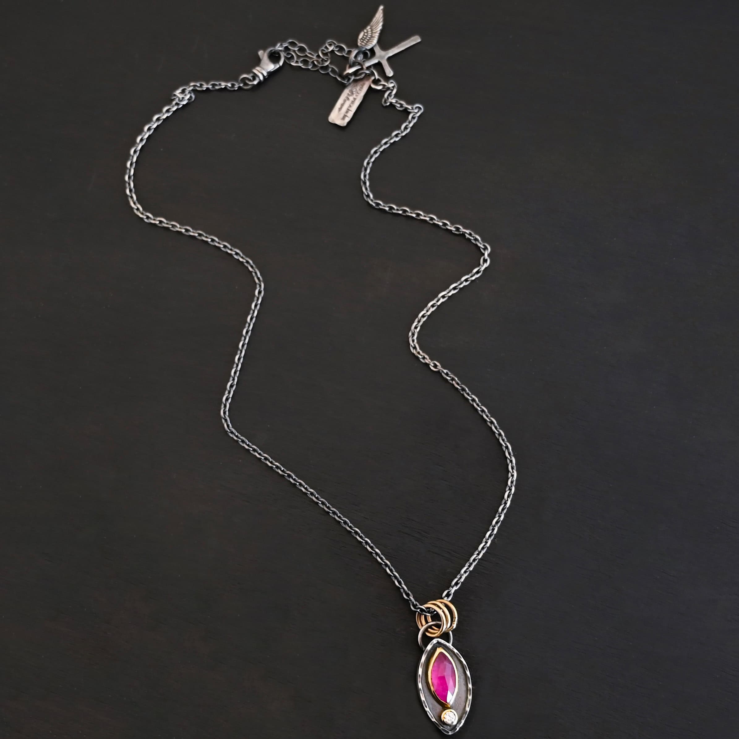 Soft Petals Necklace New Photo of the "Soft Petals" Necklace. Made out of sterling silver, gold, ruby, and diamond. The bottom of the ring has a small circle diamond. Above this is a pink, marquise shaped ruby placed vertically, so that the points are pointing up and down. The stone is set in gold, and there are two layers of sterling silver beneath it. The top layer of silver goes around the edge of the bottom layer. Both layers are marquise shaped, and the top outside layer is hammered. There is a loop on the back of the necklace in which three gold jump rings go through. Going through the jump rings is the silver chain of the necklace. Necklace is laying against a dark background.