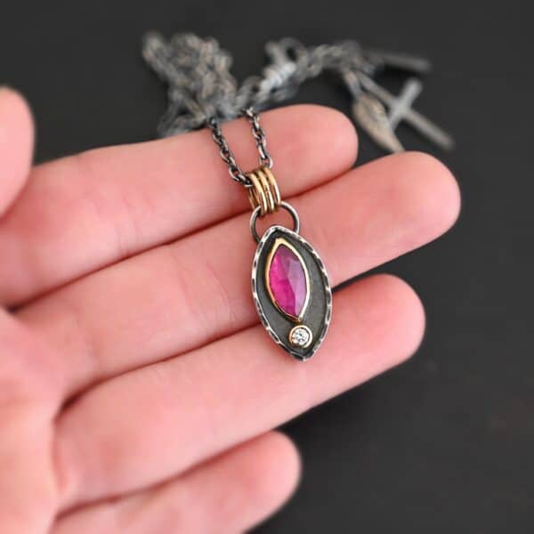 Photo of the "Soft Petals" Necklace. Made out of sterling silver, gold, ruby, and diamond. The bottom of the ring has a small circle diamond. Above this is a pink, marquise shaped ruby placed vertically, so that the points are pointing up and down. The stone is set in gold, and there are two layers of sterling silver beneath it. The top layer of silver goes around the edge of the bottom layer. Both layers are marquise shaped, and the top outside layer is hammered. There is a loop on the back of the necklace in which three gold jump rings go through. Going through the jump rings is the silver chain of the necklace. Necklace is laying against the fingertips of a hand to demonstrate size.