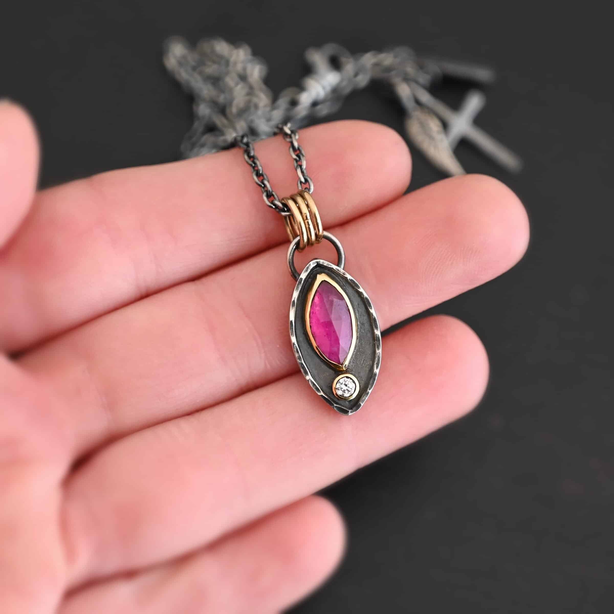 Soft Petals Necklace New Photo of the "Soft Petals" Necklace. Made out of sterling silver, gold, ruby, and diamond. The bottom of the ring has a small circle diamond. Above this is a pink, marquise shaped ruby placed vertically, so that the points are pointing up and down. The stone is set in gold, and there are two layers of sterling silver beneath it. The top layer of silver goes around the edge of the bottom layer. Both layers are marquise shaped, and the top outside layer is hammered. There is a loop on the back of the necklace in which three gold jump rings go through. Going through the jump rings is the silver chain of the necklace. Necklace is laying against the fingertips of a hand to demonstrate size.