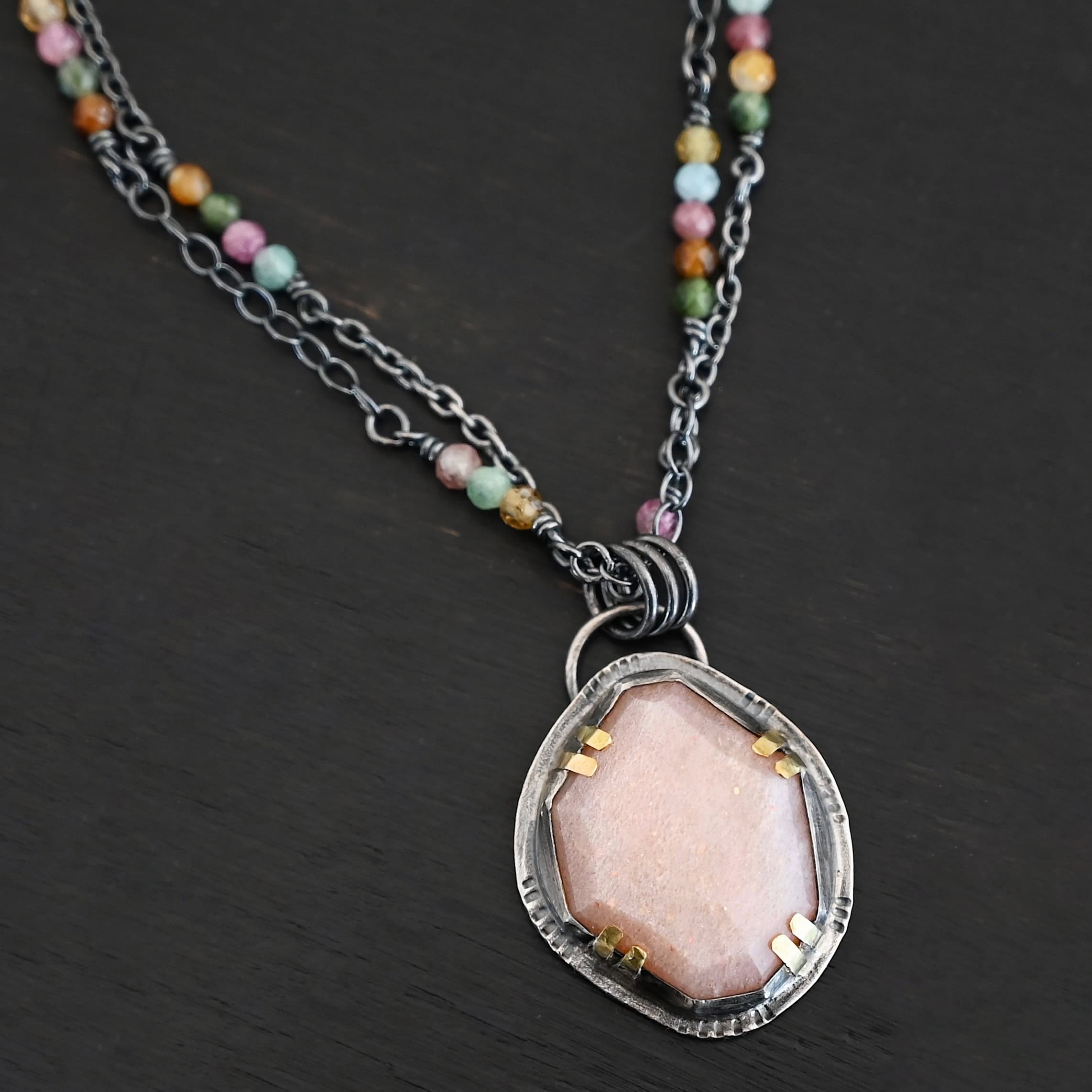 Peach Tree Necklace New Photo of the "Peach Tree" Necklace. Made out of sterling silver, gold, peach moonstone, and tourmaline. There is a rose cut, octagon shaped peach moonstone in the center with eight gold prongs holding the stone in. The prongs are four groups of two in each corner of the ring. The silver around the stone has hammered marks in groups scattered around it as well. There is a loop on the back of the pendant with three silver jump rings going through it. Going through the jump rings is the chain of the necklace. The chain of the necklace is two separate chains that meet at the back to make one chain for the necklace. Each chain has multiple tourmaline beads on it. Necklace is lying against a dark background.