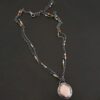Photo of the "Peach Tree" Necklace. Made out of sterling silver, gold, peach moonstone, and tourmaline. There is a rose cut, octagon shaped peach moonstone in the center with eight gold prongs holding the stone in. The prongs are four groups of two in each corner of the ring. The silver around the stone has hammered marks in groups scattered around it as well. There is a loop on the back of the pendant with three silver jump rings going through it. Going through the jump rings is the chain of the necklace. The chain of the necklace is two separate chains that meet at the back to make one chain for the necklace. Each chain has multiple tourmaline beads on it. Necklace is lying against a dark background.