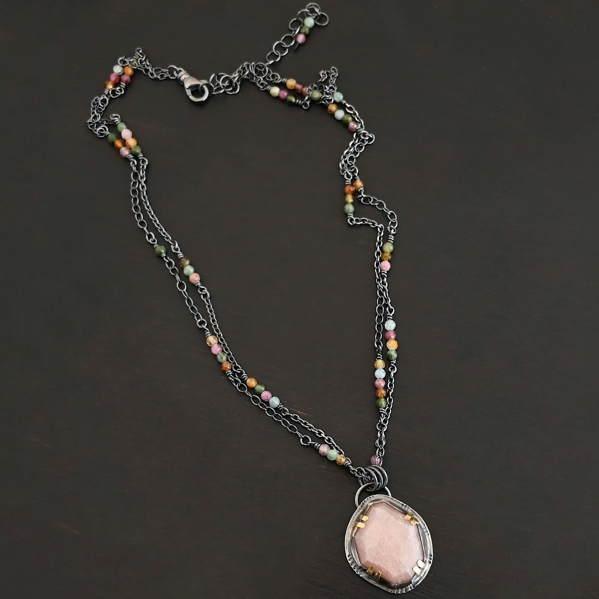 Peach Tree Necklace New Photo of the "Peach Tree" Necklace. Made out of sterling silver, gold, peach moonstone, and tourmaline. There is a rose cut, octagon shaped peach moonstone in the center with eight gold prongs holding the stone in. The prongs are four groups of two in each corner of the ring. The silver around the stone has hammered marks in groups scattered around it as well. There is a loop on the back of the pendant with three silver jump rings going through it. Going through the jump rings is the chain of the necklace. The chain of the necklace is two separate chains that meet at the back to make one chain for the necklace. Each chain has multiple tourmaline beads on it. Necklace is lying against a dark background.