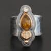 Photo of the "Reach For The Sun" Ring. Made out of sterling silver, gold, tourmaline, rutilated quartz, and lab created diamond. The lab created diamond is at the top, on the tip of the pear shaped tourmaline. The tourmaline is a dark brown color with golden flecks. There is a small circular rutilated quartz beneath the tourmaline. On the bottom of the rutilated quartz is a gold ball. Each stone is set in gold, and there are two layers of sterling silver beneath the stones. The top layer is smaller than the bottom layer, so that the bottom layer sticks out. The band of the ring is thick and hammered and oxidized. Ring is sitting against a dark background