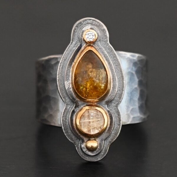 Photo of the "Reach For The Sun" Ring. Made out of sterling silver, gold, tourmaline, rutilated quartz, and lab created diamond. The lab created diamond is at the top, on the tip of the pear shaped tourmaline. The tourmaline is a dark brown color with golden flecks. There is a small circular rutilated quartz beneath the tourmaline. On the bottom of the rutilated quartz is a gold ball. Each stone is set in gold, and there are two layers of sterling silver beneath the stones. The top layer is smaller than the bottom layer, so that the bottom layer sticks out. The band of the ring is thick and hammered and oxidized. Ring is sitting against a dark background