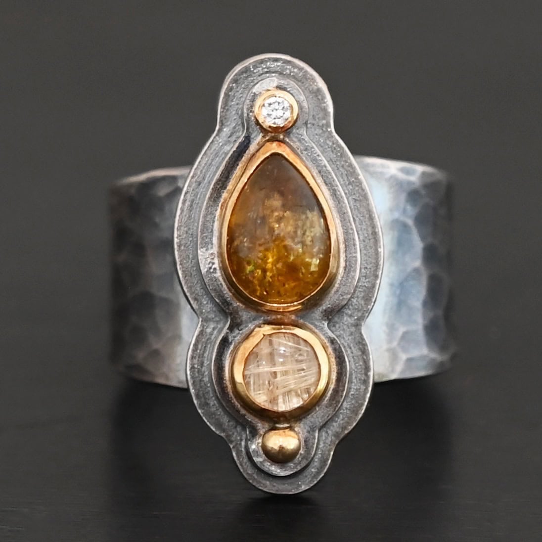 Reach For The Sun Ring New Photo of the "Reach For The Sun" Ring. Made out of sterling silver, gold, tourmaline, rutilated quartz, and lab created diamond. The lab created diamond is at the top, on the tip of the pear shaped tourmaline. The tourmaline is a dark brown color with golden flecks. There is a small circular rutilated quartz beneath the tourmaline. On the bottom of the rutilated quartz is a gold ball. Each stone is set in gold, and there are two layers of sterling silver beneath the stones. The top layer is smaller than the bottom layer, so that the bottom layer sticks out. The band of the ring is thick and hammered and oxidized. Ring is sitting against a dark background