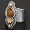 Photo of the "Reach For The Sun" Ring. Made out of sterling silver, gold, tourmaline, rutilated quartz, and lab created diamond. The lab created diamond is at the top, on the tip of the pear shaped tourmaline. The tourmaline is a dark brown color with golden flecks. There is a small circular rutilated quartz beneath the tourmaline. On the bottom of the rutilated quartz is a gold ball. Each stone is set in gold, and there are two layers of sterling silver beneath the stones. The top layer is smaller than the bottom layer, so that the bottom layer sticks out. The band of the ring is thick and hammered and oxidized. Ring is sitting against a dark background