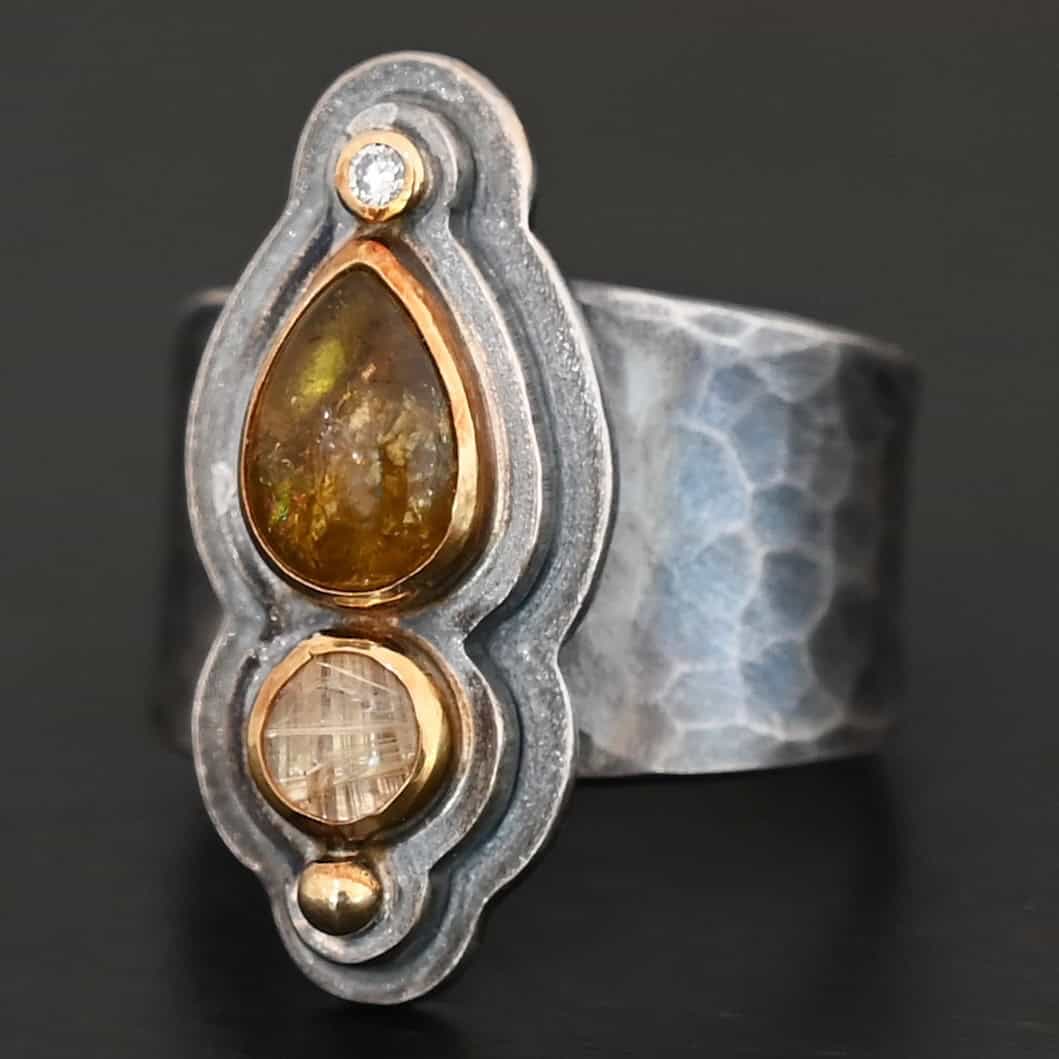 Reach For The Sun Ring New Photo of the "Reach For The Sun" Ring. Made out of sterling silver, gold, tourmaline, rutilated quartz, and lab created diamond. The lab created diamond is at the top, on the tip of the pear shaped tourmaline. The tourmaline is a dark brown color with golden flecks. There is a small circular rutilated quartz beneath the tourmaline. On the bottom of the rutilated quartz is a gold ball. Each stone is set in gold, and there are two layers of sterling silver beneath the stones. The top layer is smaller than the bottom layer, so that the bottom layer sticks out. The band of the ring is thick and hammered and oxidized. Ring is sitting against a dark background