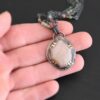 Photo of the "Peach Tree" Necklace. Made out of sterling silver, gold, peach moonstone, and tourmaline. There is a rose cut, octagon shaped peach moonstone in the center with eight gold prongs holding the stone in. The prongs are four groups of two in each corner of the ring. The silver around the stone has hammered marks in groups scattered around it as well. There is a loop on the back of the pendant with three silver jump rings going through it. Going through the jump rings is the chain of the necklace. The chain of the necklace is two separate chains that meet at the back to make one chain for the necklace. Each chain has multiple tourmaline beads on it. Necklace is lying against the fingertips of a hand to demonstrate size.