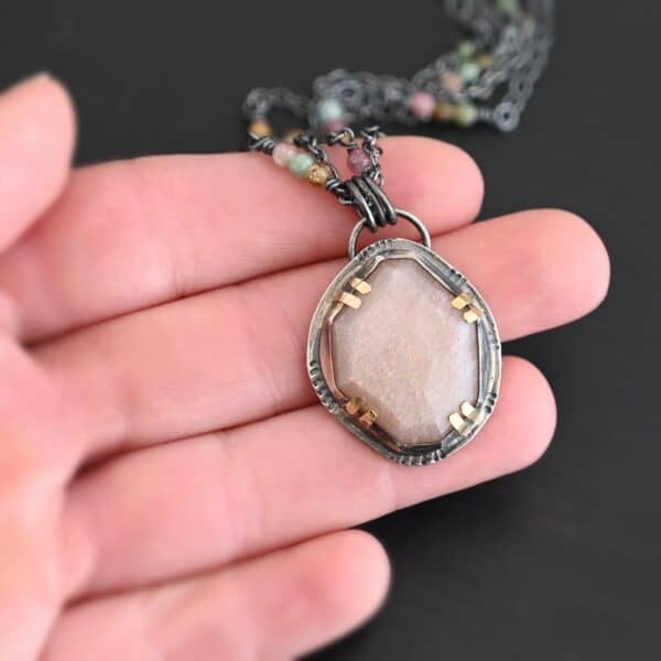 Photo of the "Peach Tree" Necklace. Made out of sterling silver, gold, peach moonstone, and tourmaline. There is a rose cut, octagon shaped peach moonstone in the center with eight gold prongs holding the stone in. The prongs are four groups of two in each corner of the ring. The silver around the stone has hammered marks in groups scattered around it as well. There is a loop on the back of the pendant with three silver jump rings going through it. Going through the jump rings is the chain of the necklace. The chain of the necklace is two separate chains that meet at the back to make one chain for the necklace. Each chain has multiple tourmaline beads on it. Necklace is lying against the fingertips of a hand to demonstrate size.