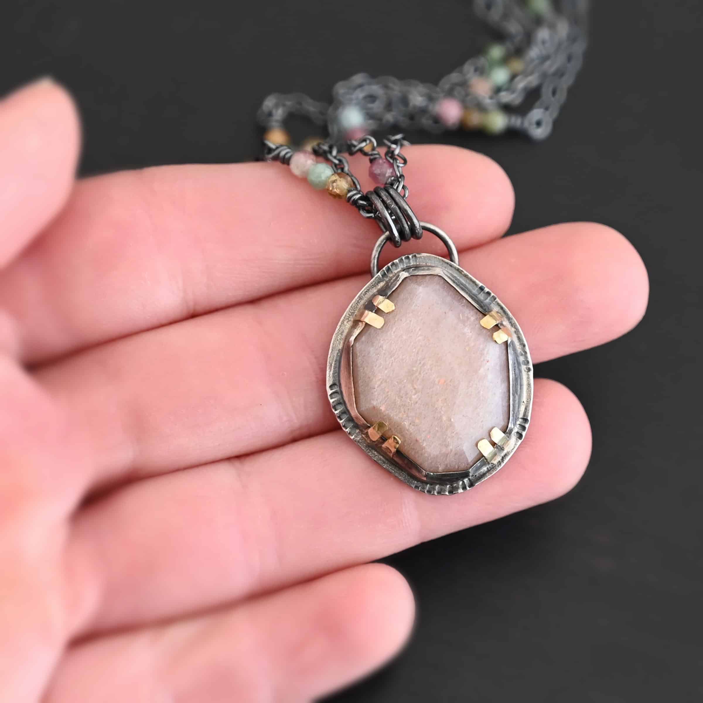 Peach Tree Necklace New Photo of the "Peach Tree" Necklace. Made out of sterling silver, gold, peach moonstone, and tourmaline. There is a rose cut, octagon shaped peach moonstone in the center with eight gold prongs holding the stone in. The prongs are four groups of two in each corner of the ring. The silver around the stone has hammered marks in groups scattered around it as well. There is a loop on the back of the pendant with three silver jump rings going through it. Going through the jump rings is the chain of the necklace. The chain of the necklace is two separate chains that meet at the back to make one chain for the necklace. Each chain has multiple tourmaline beads on it. Necklace is lying against the fingertips of a hand to demonstrate size.