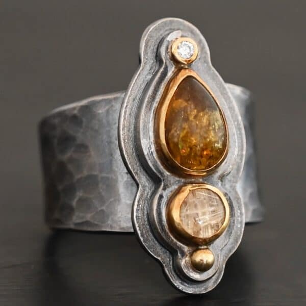 Photo of the "Reach For The Sun" Ring. Made out of sterling silver, gold, tourmaline, rutilated quartz, and lab created diamond. The lab created diamond is at the top, on the tip of the pear shaped tourmaline. The tourmaline is a dark brown color with golden flecks. There is a small circular rutilated quartz beneath the tourmaline. On the bottom of the rutilated quartz is a gold ball. Each stone is set in gold, and there are two layers of sterling silver beneath the stones. The top layer is smaller than the bottom layer, so that the bottom layer sticks out. The band of the ring is thick and hammered and oxidized. Ring is sitting against a dark background