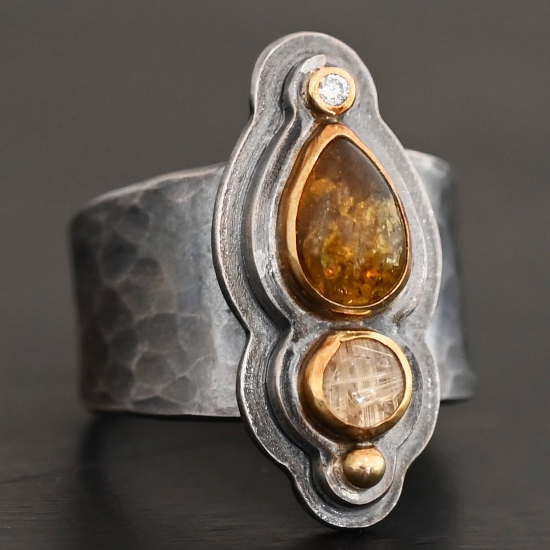 Reach For The Sun Ring New Photo of the "Reach For The Sun" Ring. Made out of sterling silver, gold, tourmaline, rutilated quartz, and lab created diamond. The lab created diamond is at the top, on the tip of the pear shaped tourmaline. The tourmaline is a dark brown color with golden flecks. There is a small circular rutilated quartz beneath the tourmaline. On the bottom of the rutilated quartz is a gold ball. Each stone is set in gold, and there are two layers of sterling silver beneath the stones. The top layer is smaller than the bottom layer, so that the bottom layer sticks out. The band of the ring is thick and hammered and oxidized. Ring is sitting against a dark background