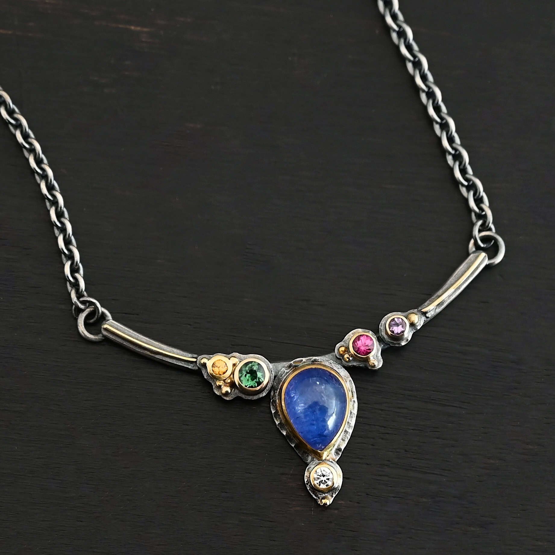 Photo of the "All In Harmony" Necklace. Made out of Sterling Silver, Gold, Tanzanite, Sapphires, Tourmaline, Amethyst, & Diamond. The center of the pendant is a pear shaped tanzanite, with the point facing down. There is a small diamond beneath the tanzanite, on the tip of the stone. On each side of the tanzanite, on the bar of the necklace that the top of the tanzanite is soldered to, are four stones, two per side. The left side on the bar has a green tourmaline and a yellow sapphire. The right side has a pink sapphire and an amethyst. There are multiple gold balls scattered around the stones on the top. There is also a thin line of gold that goes across the curved bar. Necklace is lying against a dark background.