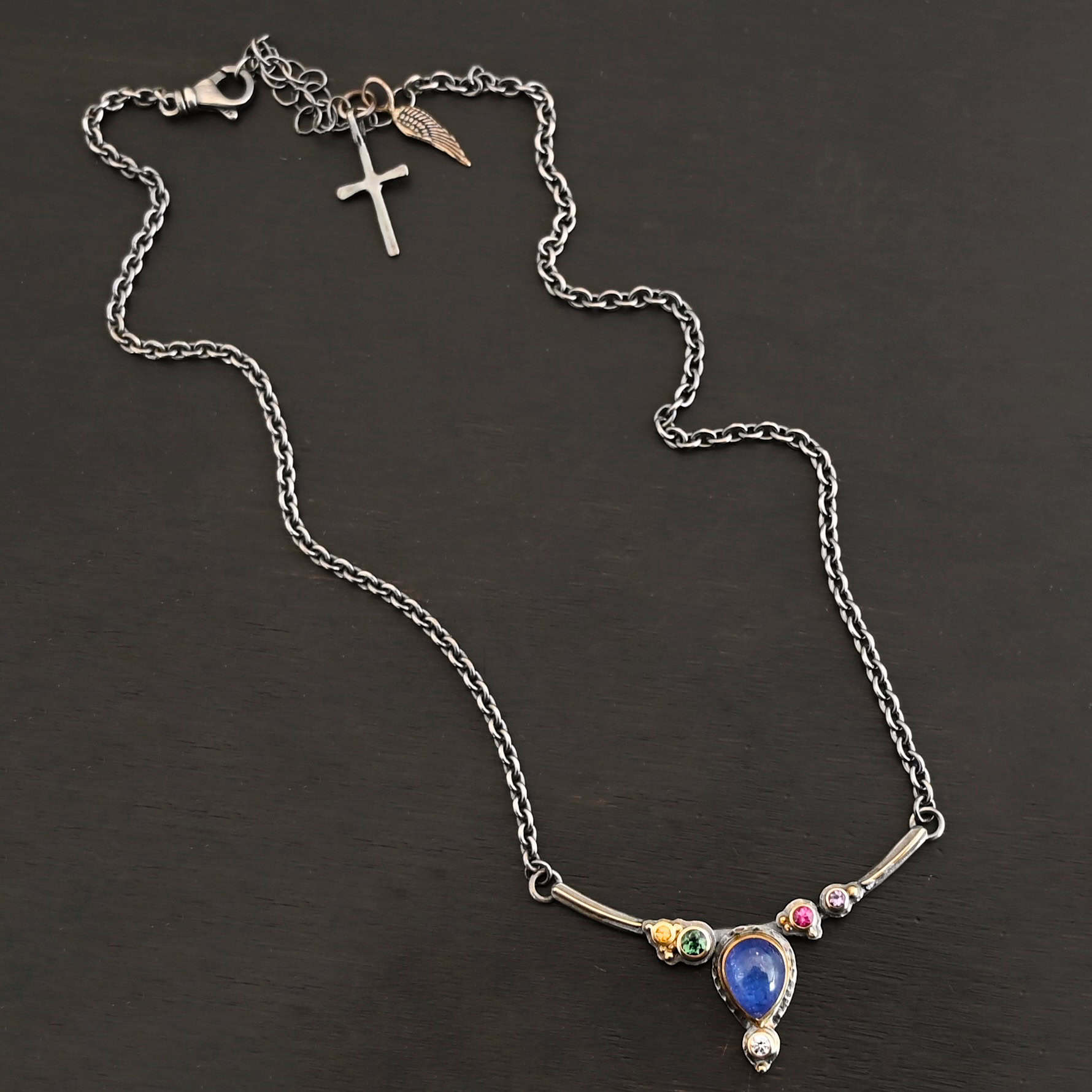 Photo of the "All In Harmony" Necklace. Made out of Sterling Silver, Gold, Tanzanite, Sapphires, Tourmaline, Amethyst, & Diamond. The center of the pendant is a pear shaped tanzanite, with the point facing down. There is a small diamond beneath the tanzanite, on the tip of the stone. On each side of the tanzanite, on the bar of the necklace that the top of the tanzanite is soldered to, are four stones, two per side. The left side on the bar has a green tourmaline and a yellow sapphire. The right side has a pink sapphire and an amethyst. There are multiple gold balls scattered around the stones on the top. There is also a thin line of gold that goes across the curved bar. Necklace is lying against a dark background.