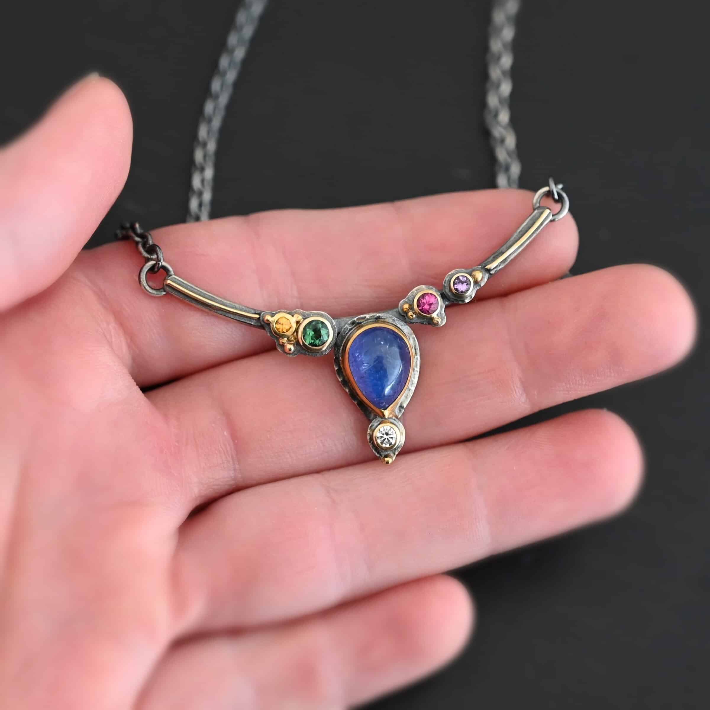 Photo of the "All In Harmony" Necklace. Made out of Sterling Silver, Gold, Tanzanite, Sapphires, Tourmaline, Amethyst, & Diamond. The center of the pendant is a pear shaped tanzanite, with the point facing down. There is a small diamond beneath the tanzanite, on the tip of the stone. On each side of the tanzanite, on the bar of the necklace that the top of the tanzanite is soldered to, are four stones, two per side. The left side on the bar has a green tourmaline and a yellow sapphire. The right side has a pink sapphire and an amethyst. There are multiple gold balls scattered around the stones on the top. There is also a thin line of gold that goes across the curved bar. Necklace is lying against the fingertips of a hand to demonstrate size.