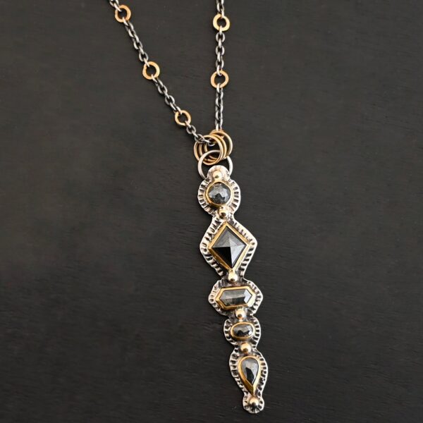 Photo of the "Nightfall" Necklace. Made out of sterling silver, gold, and diamond. There are five diamonds, all in a straight line, that create the pendant of the necklace. Each of the diamonds are different shapes, sizes, and colors. Each of these diamonds are separated by gold balls, which there are six of. The top of the pendant starts with a gold ball, then a large diamond, then a gold ball, and keeps alternating until the final diamond that has a gold ball on the end of it. There is a large loop soldered to the back of the top of the pendant with three gold jump rings attached to it. Going through the jump rings is the silver chain of the necklace. The chain of the necklace has gold loops scattered around it. Necklace is lying against a dark background.