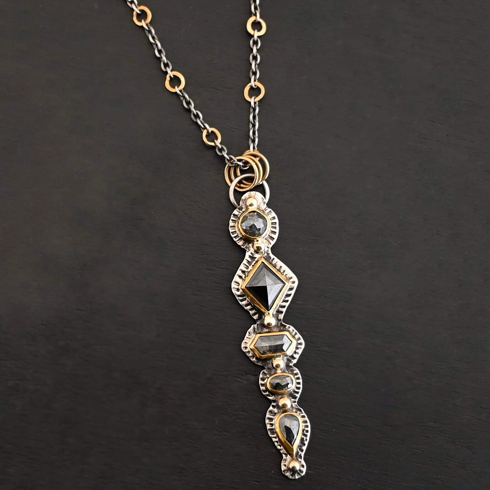 Nightfall Necklace Photo of the "Nightfall" Necklace. Made out of sterling silver, gold, and diamond. There are five diamonds, all in a straight line, that create the pendant of the necklace. Each of the diamonds are different shapes, sizes, and colors. Each of these diamonds are separated by gold balls, which there are six of. The top of the pendant starts with a gold ball, then a large diamond, then a gold ball, and keeps alternating until the final diamond that has a gold ball on the end of it. There is a large loop soldered to the back of the top of the pendant with three gold jump rings attached to it. Going through the jump rings is the silver chain of the necklace. The chain of the necklace has gold loops scattered around it. Necklace is lying against a dark background.