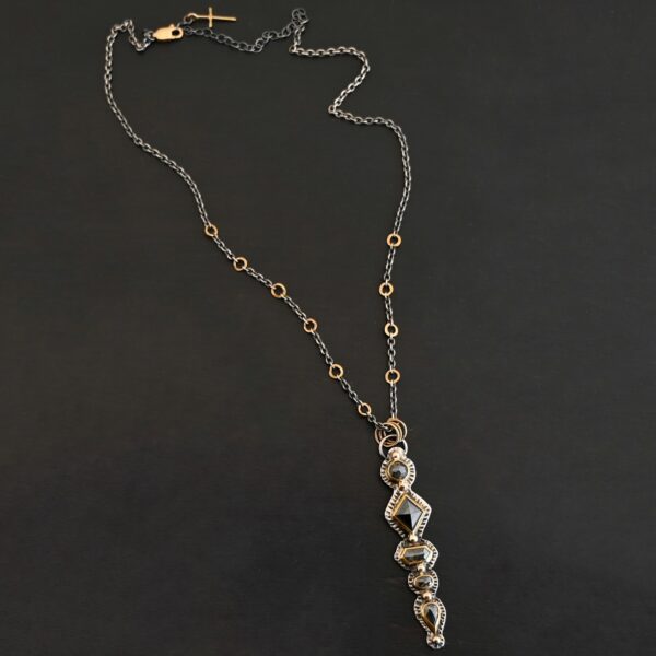 Photo of the "Nightfall" Necklace. Made out of sterling silver, gold, and diamond. There are five diamonds, all in a straight line, that create the pendant of the necklace. Each of the diamonds are different shapes, sizes, and colors. Each of these diamonds are separated by gold balls, which there are six of. The top of the pendant starts with a gold ball, then a large diamond, then a gold ball, and keeps alternating until the final diamond that has a gold ball on the end of it. There is a large loop soldered to the back of the top of the pendant with three gold jump rings attached to it. Going through the jump rings is the silver chain of the necklace. The chain of the necklace has gold loops scattered around it. Necklace is lying against a dark background.