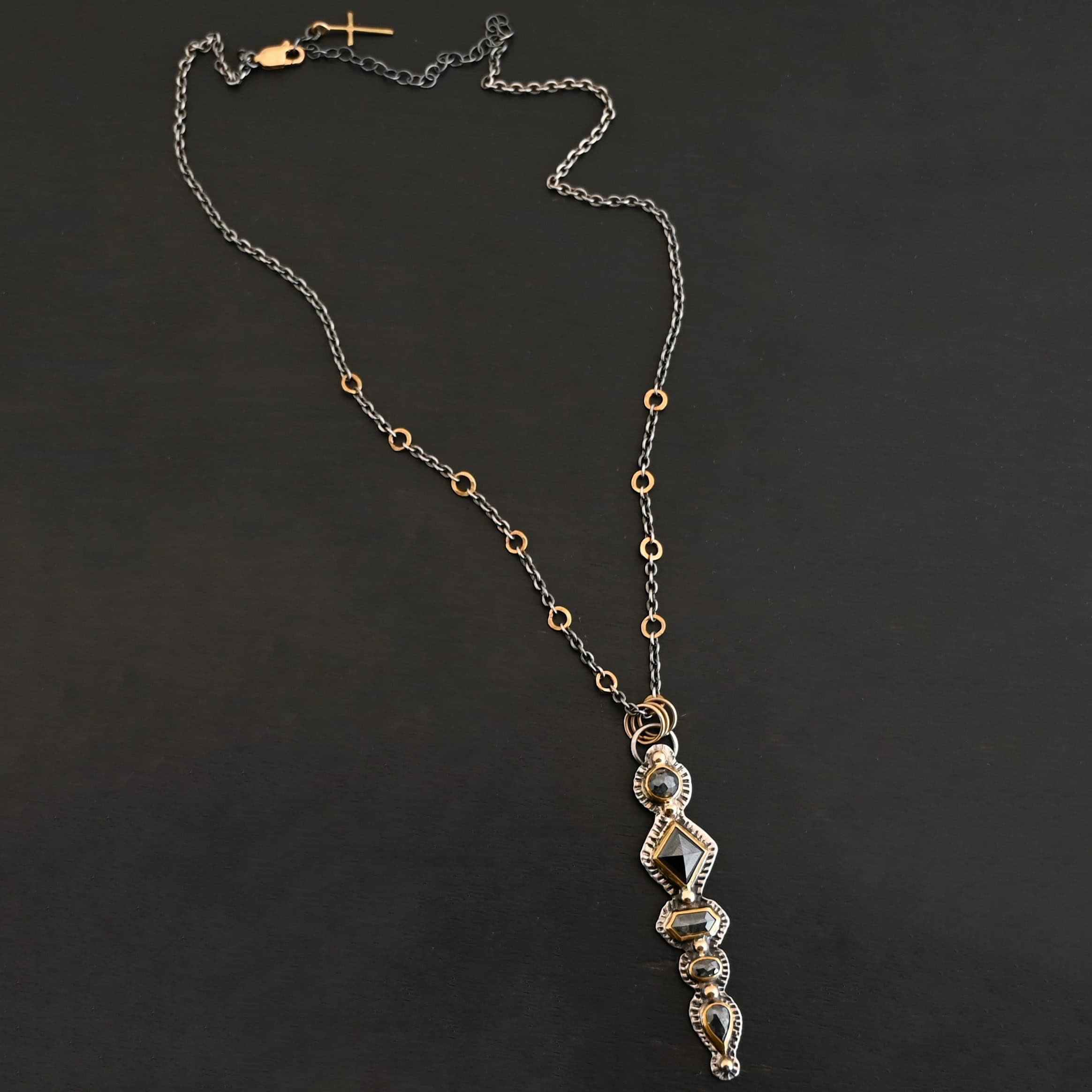 Nightfall Necklace Photo of the "Nightfall" Necklace. Made out of sterling silver, gold, and diamond. There are five diamonds, all in a straight line, that create the pendant of the necklace. Each of the diamonds are different shapes, sizes, and colors. Each of these diamonds are separated by gold balls, which there are six of. The top of the pendant starts with a gold ball, then a large diamond, then a gold ball, and keeps alternating until the final diamond that has a gold ball on the end of it. There is a large loop soldered to the back of the top of the pendant with three gold jump rings attached to it. Going through the jump rings is the silver chain of the necklace. The chain of the necklace has gold loops scattered around it. Necklace is lying against a dark background.