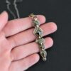 Photo of the "Nightfall" Necklace. Made out of sterling silver, gold, and diamond. There are five diamonds, all in a straight line, that create the pendant of the necklace. Each of the diamonds are different shapes, sizes, and colors. Each of these diamonds are separated by gold balls, which there are six of. The top of the pendant starts with a gold ball, then a large diamond, then a gold ball, and keeps alternating until the final diamond that has a gold ball on the end of it. There is a large loop soldered to the back of the top of the pendant with three gold jump rings attached to it. Going through the jump rings is the silver chain of the necklace. The chain of the necklace has gold loops scattered around it. Necklace is lying against the fingertips of a hand.