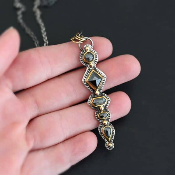 Photo of the "Nightfall" Necklace. Made out of sterling silver, gold, and diamond. There are five diamonds, all in a straight line, that create the pendant of the necklace. Each of the diamonds are different shapes, sizes, and colors. Each of these diamonds are separated by gold balls, which there are six of. The top of the pendant starts with a gold ball, then a large diamond, then a gold ball, and keeps alternating until the final diamond that has a gold ball on the end of it. There is a large loop soldered to the back of the top of the pendant with three gold jump rings attached to it. Going through the jump rings is the silver chain of the necklace. The chain of the necklace has gold loops scattered around it. Necklace is lying against the fingertips of a hand.
