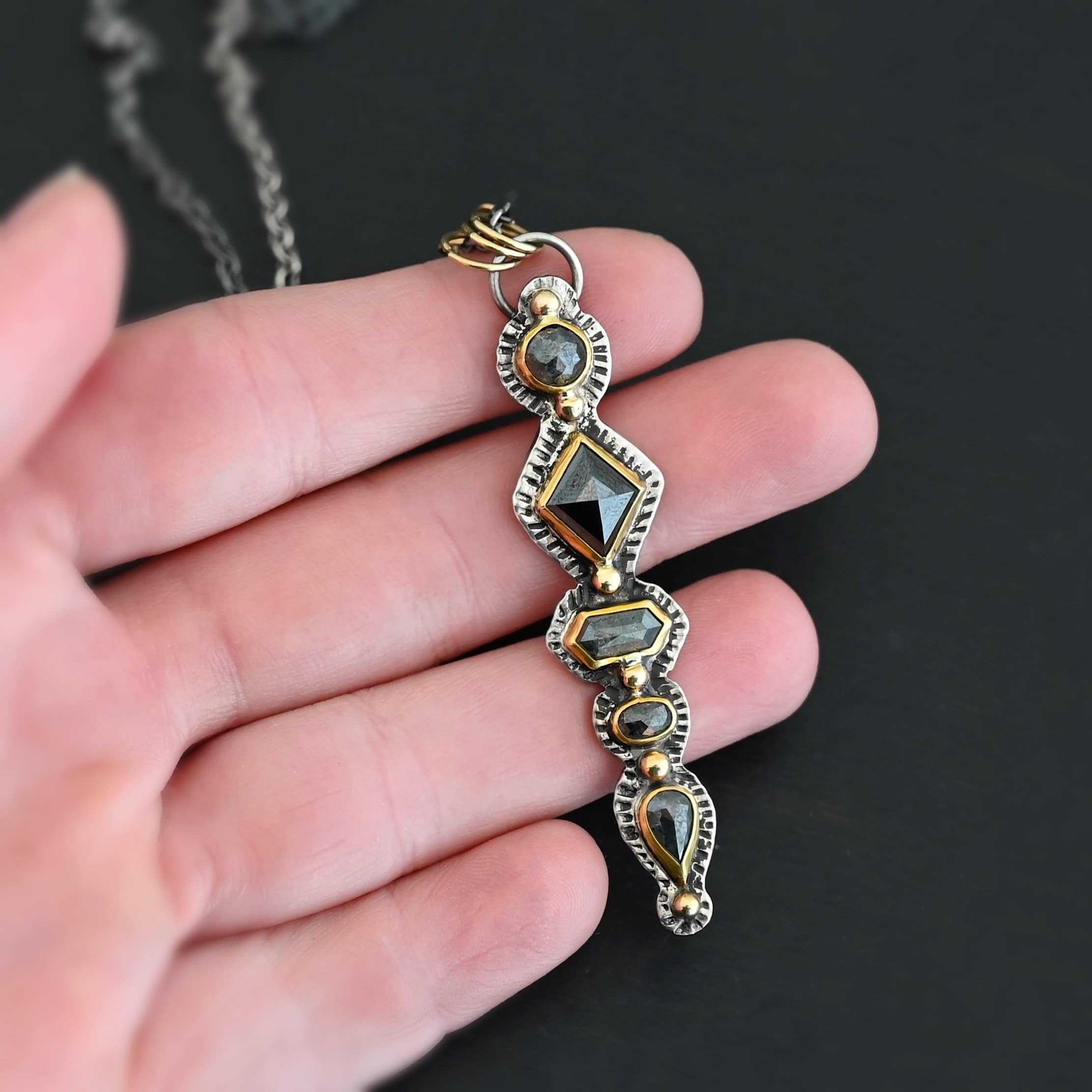 Nightfall Necklace Photo of the "Nightfall" Necklace. Made out of sterling silver, gold, and diamond. There are five diamonds, all in a straight line, that create the pendant of the necklace. Each of the diamonds are different shapes, sizes, and colors. Each of these diamonds are separated by gold balls, which there are six of. The top of the pendant starts with a gold ball, then a large diamond, then a gold ball, and keeps alternating until the final diamond that has a gold ball on the end of it. There is a large loop soldered to the back of the top of the pendant with three gold jump rings attached to it. Going through the jump rings is the silver chain of the necklace. The chain of the necklace has gold loops scattered around it. Necklace is lying against the fingertips of a hand.