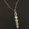 Photo of the "Daybreak" Necklace. Made out of sterling silver, gold, and diamond. There are nine diamonds, all in a straight line, that create the pendant of the necklace. Five of the diamonds are different shapes, sizes, and colors. Each of these diamonds are separated by small, circular diamonds, which there are four of. The top of the pendant starts with a large diamond, then a small one, and keeps alternating until the final diamond is a large one. There is a large loop soldered to the back of the top of the pendant with four gold jump rings attached to it. Going through the jump rings is the silver chain of the necklace. The chain of the necklace has gold loops scattered around it. Necklace is lying against a dark background.