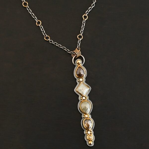 Photo of the "Daybreak" Necklace. Made out of sterling silver, gold, and diamond. There are nine diamonds, all in a straight line, that create the pendant of the necklace. Five of the diamonds are different shapes, sizes, and colors. Each of these diamonds are separated by small, circular diamonds, which there are four of. The top of the pendant starts with a large diamond, then a small one, and keeps alternating until the final diamond is a large one. There is a large loop soldered to the back of the top of the pendant with four gold jump rings attached to it. Going through the jump rings is the silver chain of the necklace. The chain of the necklace has gold loops scattered around it. Necklace is lying against a dark background.