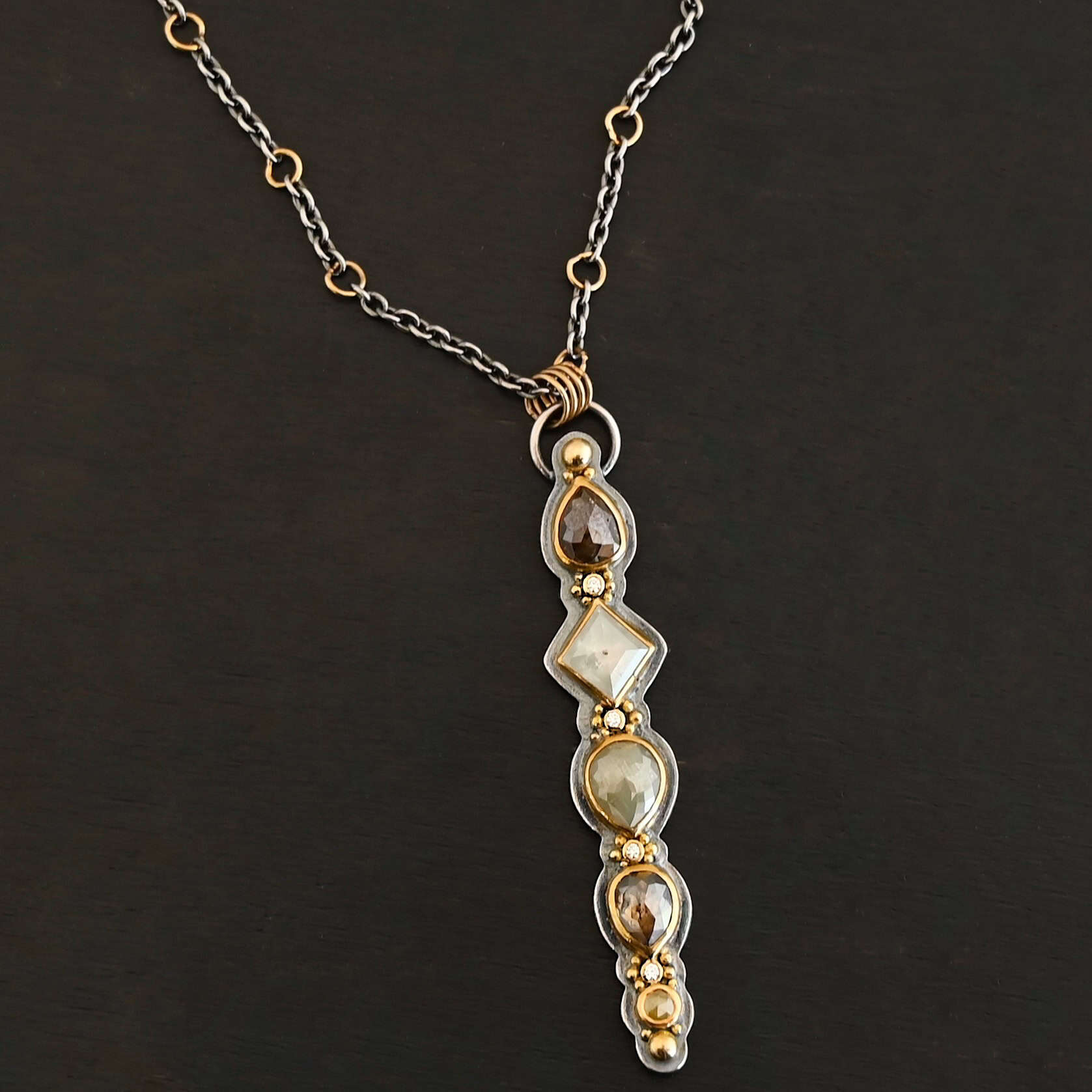 Daybreak Necklace Photo of the "Daybreak" Necklace. Made out of sterling silver, gold, and diamond. There are nine diamonds, all in a straight line, that create the pendant of the necklace. Five of the diamonds are different shapes, sizes, and colors. Each of these diamonds are separated by small, circular diamonds, which there are four of. The top of the pendant starts with a large diamond, then a small one, and keeps alternating until the final diamond is a large one. There is a large loop soldered to the back of the top of the pendant with four gold jump rings attached to it. Going through the jump rings is the silver chain of the necklace. The chain of the necklace has gold loops scattered around it. Necklace is lying against a dark background.