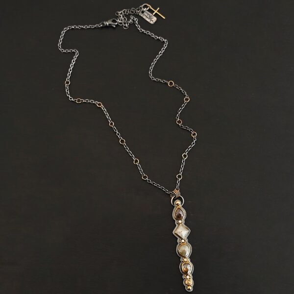 Photo of the "Daybreak" Necklace. Made out of sterling silver, gold, and diamond. There are nine diamonds, all in a straight line, that create the pendant of the necklace. Five of the diamonds are different shapes, sizes, and colors. Each of these diamonds are separated by small, circular diamonds, which there are four of. The top of the pendant starts with a large diamond, then a small one, and keeps alternating until the final diamond is a large one. There is a large loop soldered to the back of the top of the pendant with four gold jump rings attached to it. Going through the jump rings is the silver chain of the necklace. The chain of the necklace has gold loops scattered around it. Necklace is lying against a dark background.